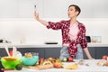 Pretty woman taking selfie or making a video call using her smartphone holding it in outstretched arm while cooking Royalty Free Stock Photo