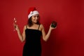 Pretty woman wearing Santa hat and black velvet dress holds champagne flute with sparkling wine and gift box, smiles toothy smile Royalty Free Stock Photo