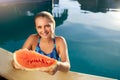 Pretty tanned slim young woman holds slice red watermelon over blue pool, relaxing on tropical island in resort, eating Royalty Free Stock Photo