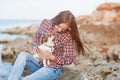 Pretty young adult woman wearing plaid shirt and jeans sitting on sea coast holding her little chihuahua dog Royalty Free Stock Photo