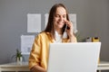 Pretty young adult woman working on laptop at her workplace in office, talking with client with charming smile, being pleased and Royalty Free Stock Photo