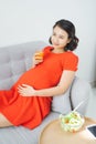 Pretty young Asian pregnant woman eating salad and drinking milk when sitting on sofa Royalty Free Stock Photo