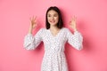 Pretty young asian woman in dress showing okay sign, praising and showing approval, looking satisfied, standing against Royalty Free Stock Photo