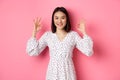 Pretty young asian woman in dress showing okay sign, praising and showing approval, looking satisfied, standing against Royalty Free Stock Photo