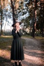 Pretty young beautiful woman in dark dress and witch hat standing in middle of autumn woods or park. Halloween party Royalty Free Stock Photo