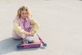 Pretty young blond curly haired hipster woman sits on the ground smiling, legs crossed, pink scooter in front of her Royalty Free Stock Photo