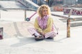 Pretty young blond curly haired woman sitting on the grey ground and looking away, smiling, legs crossed, a scooter on Royalty Free Stock Photo