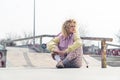 Pretty young blond ruddy hipster woman sits on the skateboard ground smiling, legs crossed, looking away pink scooter on Royalty Free Stock Photo