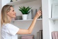 Pretty young blond woman at home is choosing a book to read in the library and smiling. wearing white shirt Royalty Free Stock Photo