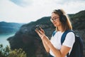 Pretty young blonde girl with glasses and backpack listens to her favorite music in wireless headphones and uses smartphone while Royalty Free Stock Photo