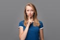 Pretty young blonde woman keeps finger on lips, making hush gesture. Shh, silence concept. Studio shot, gray background Royalty Free Stock Photo