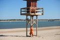 Pretty young blonde woman in orange dress leaning on the wooden lifeguard tower on the beach. The woman is nostalgic and sad. In