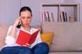 Pretty young brunette woman bored reading a book sitting on the sofa at home. Royalty Free Stock Photo