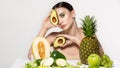 Pretty young brunette woman covering eye with fresh ripe green avocado, sits by the table with fruits and vegetabels Royalty Free Stock Photo