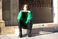 pretty young brunette woman with curly hair and green woollen coat is sitting on a step at the entrance of a building in seville,