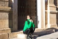 pretty young brunette woman with curly hair and green woollen coat is sitting on a step at the entrance of a building in seville,