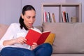 Pretty young brunette woman surprised reading a book sitting on the sofa at home.