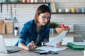 Pretty young business woman working with computer while consulting some invoices and documents in the kitchen at home Royalty Free Stock Photo