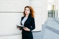 Pretty young businesswoman wearing black jacket, skirt and white blouse, holding her pocket book with pen, writing notes or lookin