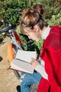 Pretty Young Caucasian Girl in Red Jacket Reading a Book Sitting on a Park Bench, with the Bicycle Beside Royalty Free Stock Photo