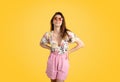 Pretty young caucasian lady wearing trendy summer outfit and colorful sunglasses, posing on yellow studio background Royalty Free Stock Photo