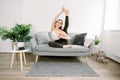 Pretty young Caucasian yoga fit woman sitting on gray sofa and posing to camera, stretching leg up, smiling and holding