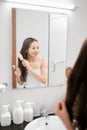 Pretty young female looking in mirror while brushing hair standing in bathroom