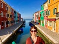 A pretty young female tourist poses on a bridge for a photo with the old colourful historic homes and canal of Burano, Italy in th