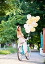 Pretty young female in white dress holding ballons and looking back while riding blue bike in a park. Royalty Free Stock Photo