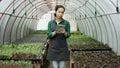 Pretty young ferm worker stands near the seedlings and texting in tablet computer inside greenhouse