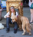 Pretty young girl with braces and long red hair with steampunk goggles sits on curb with labradoodle dog dressed in leather coat a