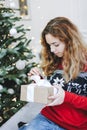 Pretty young girl opening presents near Christmas tree. Girl in red knitted sweater holds gift box Royalty Free Stock Photo