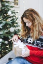 Pretty young girl opening presents near Christmas tree. Girl in red knitted sweater holds gift box Royalty Free Stock Photo