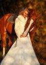 pretty young girl in wedding dress posing with her bright red horse in forest at sunny autumn day