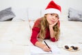 Pretty young girl writing letter for Santa Claus. Child in red santa hat lying on bed  and fhinking and writing christmas letter. Royalty Free Stock Photo
