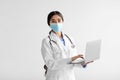 Pretty young hindu lady doctor in white coat with stethoscope in protective mask works on laptop on light background Royalty Free Stock Photo