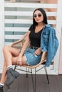 Pretty young hipster woman with long hair in stylish denim clothes in sunglasses in leather autumn boots sits on a vintage chair Royalty Free Stock Photo
