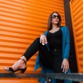 Pretty young hipster woman in stylish clothes in trendy sunglasses in trendy sandals poses near the modern orange metal wall