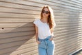 Pretty young hipster woman in stylish sunglasses in trendy white t-shirt in vintage jeans poses near a wooden wall Royalty Free Stock Photo