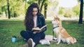 Pretty young lady is reading book in the park and patting cute dog shiba inu breed, well-bred pet is sitting on the