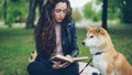 Pretty young lady is reading book in the park and patting cute dog shiba inu breed, well-bred pet is sitting on the