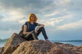 Pretty young lady with red hair sitting on stone near bay shore in summer evening. Tourist on the beautiful landscape background. Royalty Free Stock Photo