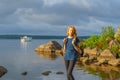 Pretty young lady with red hair standing on stone near bay shore in summer evening. Tourist on the beautiful landscape background Royalty Free Stock Photo