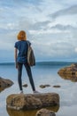 Pretty young lady with red hair standing on stone near bay shore in summer evening. Tourist on the beautiful landscape background Royalty Free Stock Photo