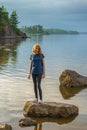 Pretty young lady with red hair standing on stone near bay shore in summer evening. Tourist on the beautiful landscape background Royalty Free Stock Photo