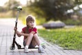 Pretty young long-haired thoughtful blond child girl in shorts and t-shirt sits on scooter on empty sunny suburb road blurred