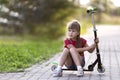 Pretty young long-haired thoughtful blond child girl in shorts and t-shirt sits on scooter on empty sunny suburb road blurred