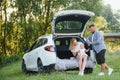 Pretty young married couple and their daughter are resting in the nature. The woman and girl are sitting on open car boot. The man Royalty Free Stock Photo