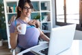 Pretty young mother with her baby in sling drinking coffee while working with laptop at home Royalty Free Stock Photo