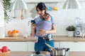 Pretty young mother with little baby in sling using her mobile phone in the kitchen at home Royalty Free Stock Photo